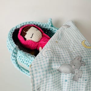 May include: A crocheted light blue basket with a pink crocheted doll inside. The doll is wearing a red hooded cloak. A light blue and white checkered blanket with a moon, stars, and bunnies printed on it is draped over the basket.