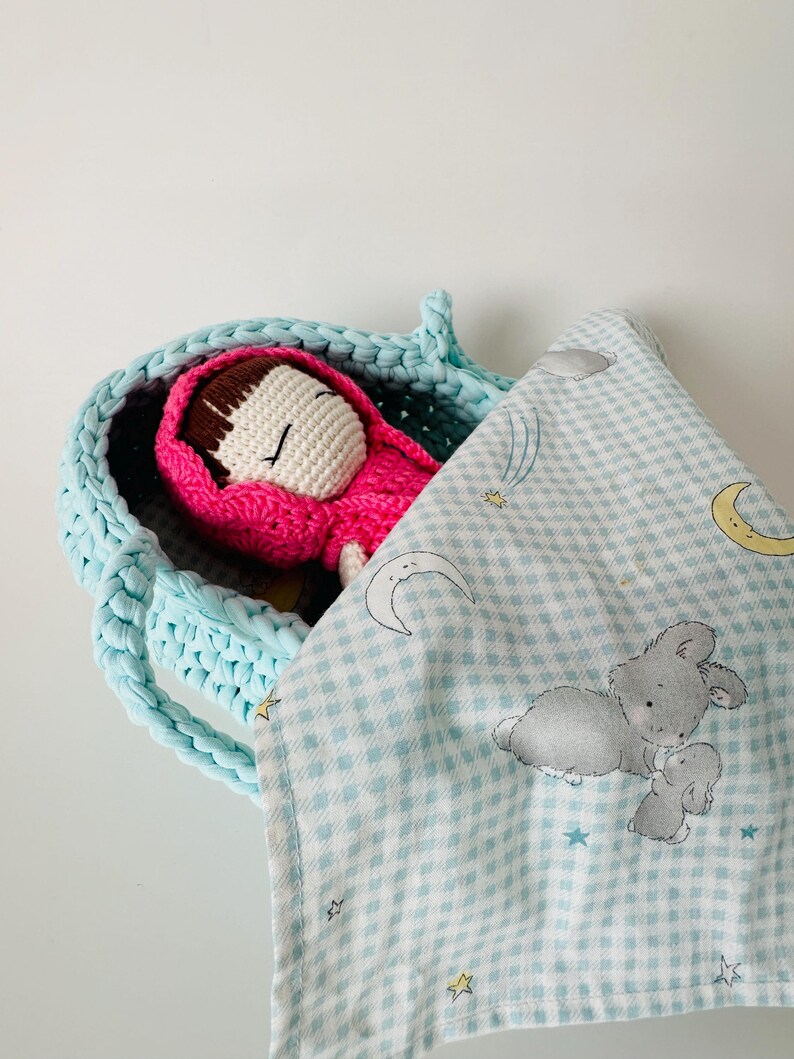 May include: A crocheted light blue basket with a pink crocheted doll inside. The doll is wearing a red hooded cloak. A light blue and white checkered blanket with a moon, stars, and bunnies printed on it is draped over the basket.