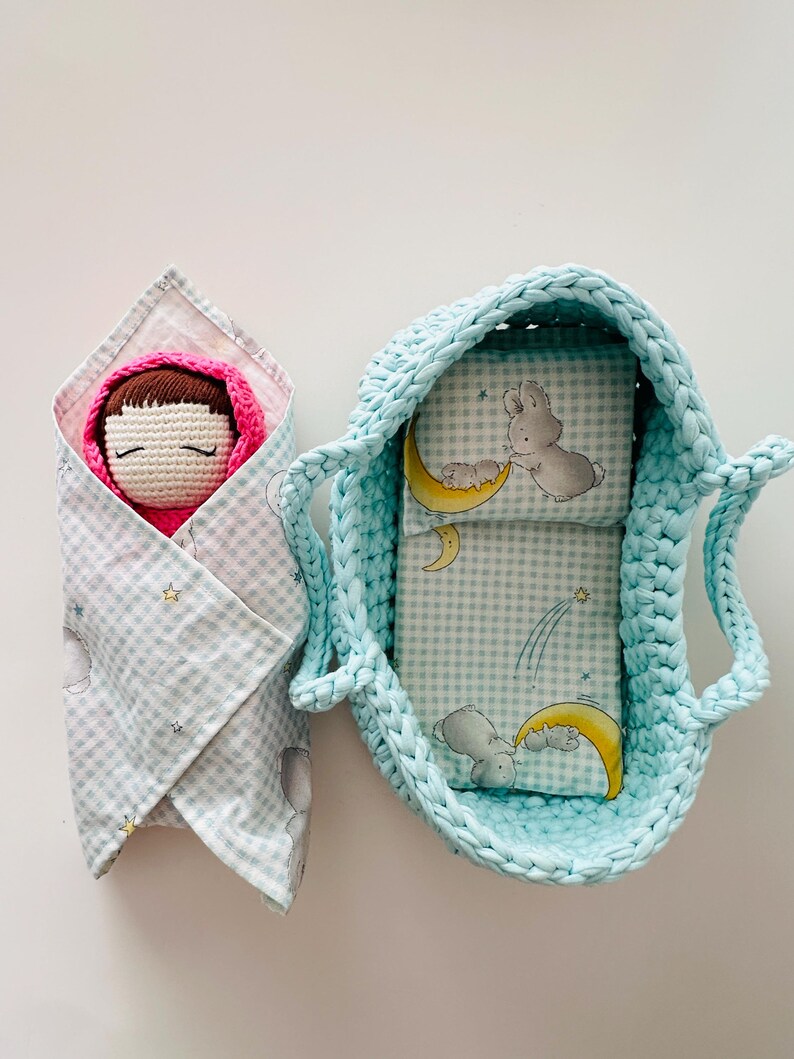 May include: A crocheted baby doll wrapped in a blue and white gingham blanket with stars and moons. The doll is in a blue crocheted basket with a blue and white gingham lining with a moon and bunny rabbit design.