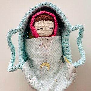 May include: A crocheted doll with brown hair and closed eyes is sleeping in a light blue crocheted basket lined with a light blue and white fabric with a moon and star pattern.