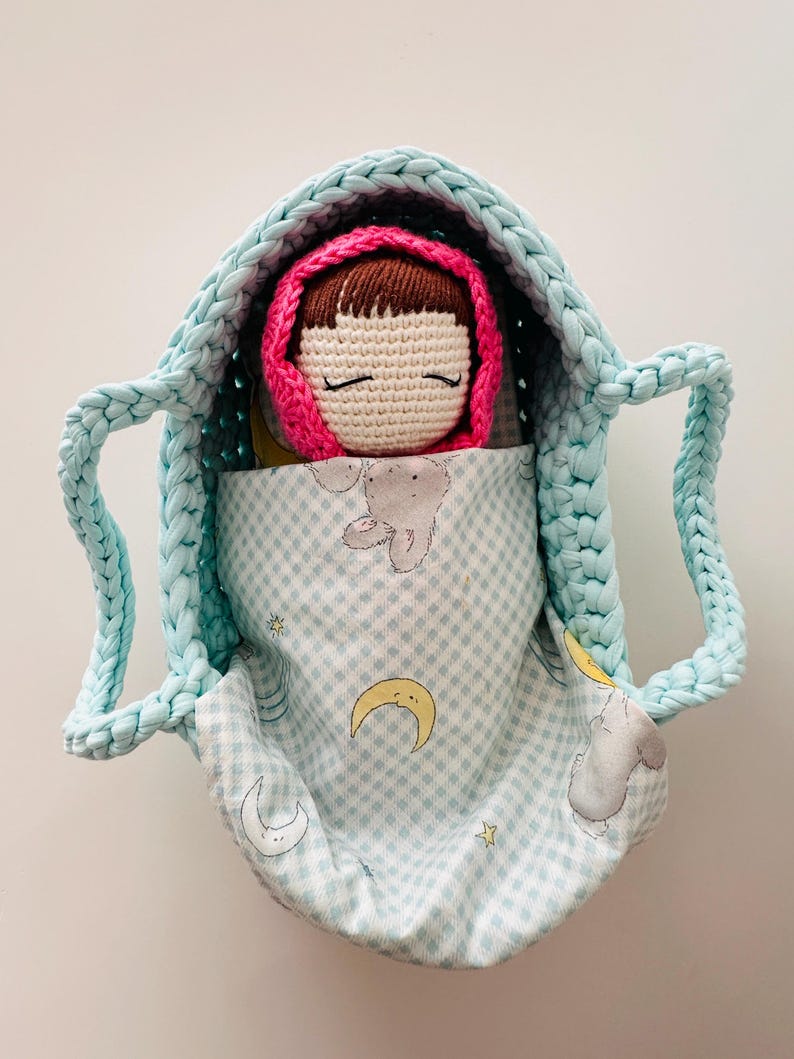 May include: A crocheted doll with brown hair and closed eyes is sleeping in a light blue crocheted basket lined with a light blue and white fabric with a moon and star pattern.