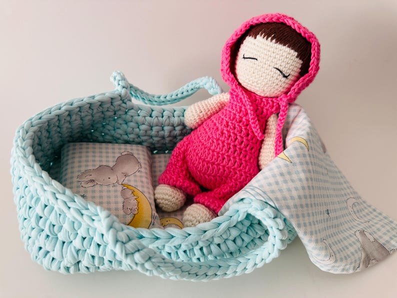 May include: A crocheted doll with a pink dress and a white hood is lying in a light blue crocheted basket with a white and blue gingham blanket. The doll has brown hair and closed eyes.