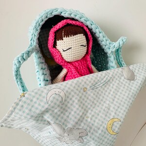 May include: A crocheted doll with brown hair and closed eyes is sleeping in a light blue crocheted basket. The basket is lined with a light blue and white checkered fabric with a moon and stars pattern.
