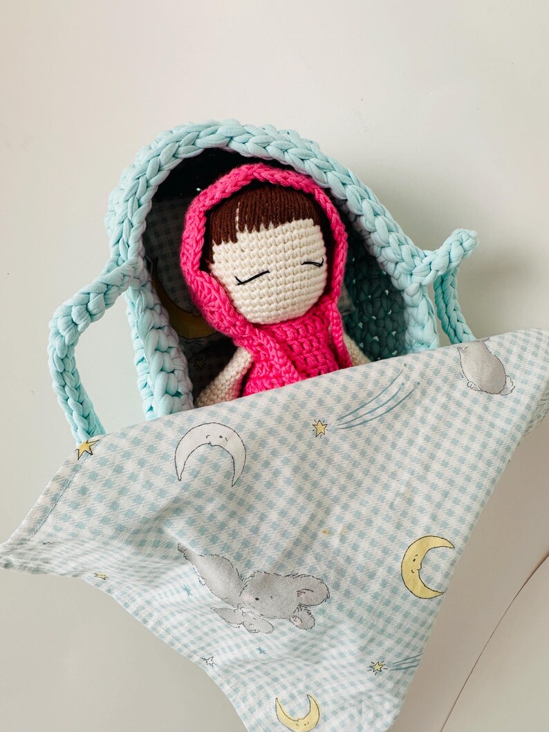 May include: A crocheted doll with brown hair and closed eyes is sleeping in a light blue crocheted basket. The basket is lined with a light blue and white checkered fabric with a moon and stars pattern.