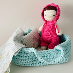 May include: A crocheted doll with a pink dress and a hood, lying in a light blue crocheted basket with a white and blue checkered blanket and a pillow.