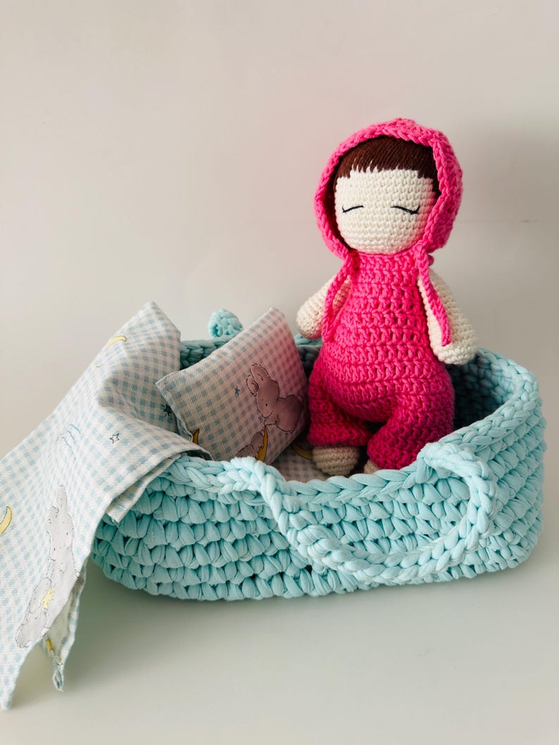 May include: A crocheted doll with a pink dress and a hood, lying in a light blue crocheted basket with a white and blue checkered blanket and a pillow.