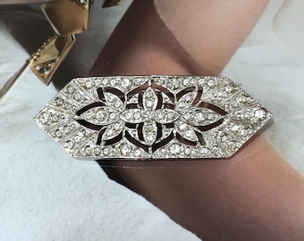 May include: A silver-toned, Art Deco-style brooch. The rectangular brooch features a floral design with clear, sparkling rhinestones. The brooch has a geometric shape with pointed ends and a central floral motif.