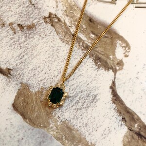 Può includere: Una collana con catena dorata con un ciondolo di pietra preziosa verde. Il ciondolo è circondato da piccole pietre preziose trasparenti.