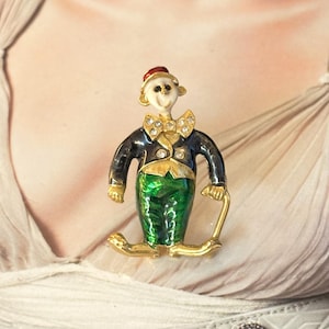 Könnte beinhalten: Eine goldfarbene Metallbrosche in Form einer Clownfigur. Der Clown trägt einen roten Hut, ein weißes Gesicht, eine goldene Fliege mit klaren Strasssteinen, eine dunkelblaue Jacke mit goldenen Akzenten und eine grüne Hose. Die Figur hält einen goldenen Stock.