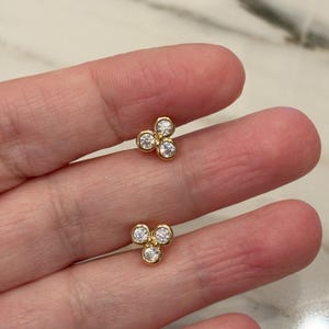 May include: Two gold stud earrings, each with three clear, round gemstones. The earrings have a gold-tone metal setting, forming a delicate floral or clover shape. The gemstones sparkle against the gold. The earrings are small and elegant.