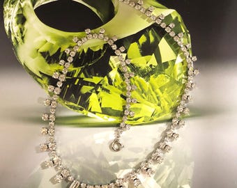 Peut inclure: Un collier argenté étincelant orné de cristaux clairs, rectangulaires et ronds. Le collier est présenté devant une grande sculpture en verre vert abstraite. Le collier a un design délicat avec une fermeture à fermoir.
