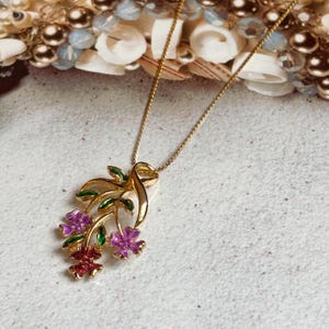 Peut inclure: Un collier doré avec un pendentif floral. Le pendentif présente des fleurs violettes et rouges avec des feuilles vertes. Le collier est posé sur une surface claire avec des éléments décoratifs en arrière-plan.
