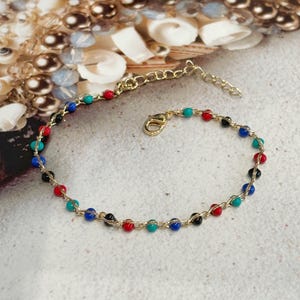 Puede incluir: Un brazalete de cadena dorada con cuentas de colores. Las cuentas son rojas, azules, negras y turquesa.