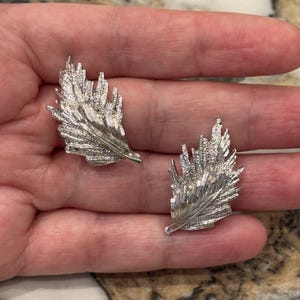 Peut inclure: Une paire de boucles d'oreilles argentées en forme de feuille. Chaque boucle d'oreille a une surface texturée avec des détails complexes, ressemblant à une plume. Les boucles d'oreilles sont présentées sur un fond neutre.