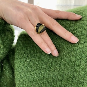 Pode incluir: Um anel de ouro com uma grande gema triangular preta e dourada. O anel é usado em um dedo.