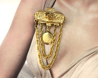 May include: A gold-toned brooch with a floral design and a dangling chain. The brooch features a rectangular centrepiece with a raised rose and intricate scrollwork. Three chains hang from the bottom of the centrepiece, each ending in a small round charm.