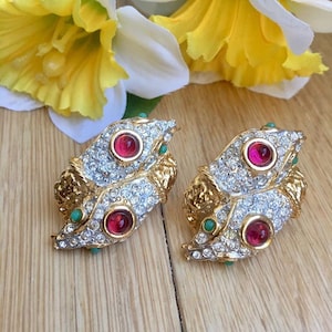 May include: A pair of vintage clip-on earrings. Each earring features a gold-tone base with a silver-tone, crystal-studded top. The earrings have a red center stone and small green accents. The earrings are set against a wooden surface.