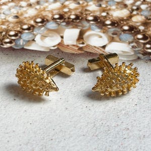 May include: A pair of gold cufflinks with a textured, spiky design. The cufflinks are in the shape of a small, round object with a pointed end.