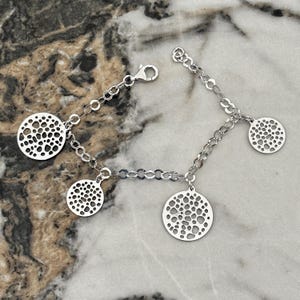 Peut inclure: Un bracelet à chaîne argentée orné de quatre breloques circulaires, chacune présentant un motif découpé de trous irréguliers. Le bracelet comprend un fermoir mousqueton et une extension de chaîne réglable pour un ajustement sûr.
