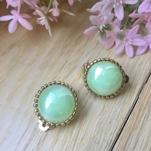 May include: A pair of clip-on earrings featuring a light green cabochon stone set in a gold-toned metal frame.
