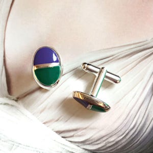 Può includere: Un paio di gemelli d'argento con un design in smalto verde e viola. I gemelli sono di forma ovale e hanno una finitura liscia e lucida.