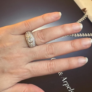 Può includere: Un anello d'argento con una pietra centrale a forma ovale di diamante, circondata da diamanti più piccoli. L'anello è su una mano, con uno sfondo scuro. Il design dell'anello è uno stile classico ed elegante, adatto alle occasioni speciali.