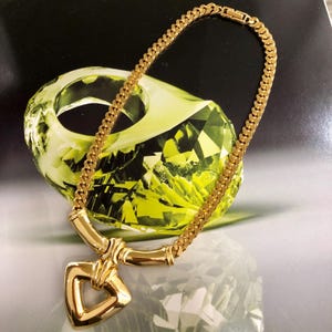 Op de afbeelding: Een gouden ketting met een grote, geometrische hanger. De hanger heeft de vorm van een hart met een uitsparing in het midden. De ketting is gemaakt van een dikke, platte ketting.