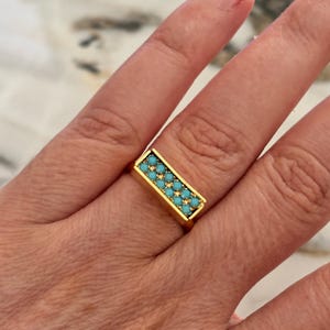 Op de afbeelding: Een goudkleurige ring met een rechthoekige zetting in de vorm van een balk. Het bovenoppervlak van de balk is ingelegd met twee parallelle rijen kleine, ronde, helder turquoise stenen, wat een gestructureerd, rasterachtig patroon op het sieraad vormt.