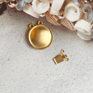 Puede incluir: Cierre de metal dorado con un disco redondo y plano y un cierre rectangular más pequeño. Los cierres están sobre una superficie blanca texturizada.