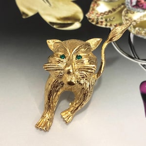 Peut inclure: Broche chat dorée avec des yeux en pierres précieuses vertes. Le chat est représenté en position de marche, la queue levée. La broche a une surface texturée, lui donnant un aspect détaillé. Bijou de style vintage.