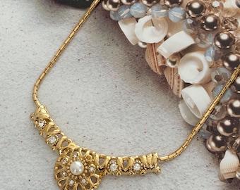 May include: A gold necklace with a delicate chain and a large, ornate pendant. The pendant features a circular design with a pearl in the center and small rhinestones around it.