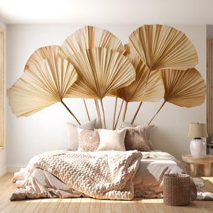 Op de afbeelding: Een slaapkamer met een bed bedekt met een beige gebreide deken en kussens. Grote, waaiervormige gedroogde palmbladeren zijn boven het bed aan de muur bevestigd. Een kleine houten bijzettafel met een lamp en een rieten mand maken de inrichting compleet.