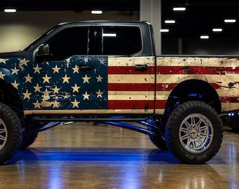 Pode incluir: Uma pick-up modificada com um design desgastado da bandeira americana na lateral. O veículo apresenta um campo de estrelas azul e branco na frente e listras vermelhas e brancas na traseira. Possui rodas cromadas grandes e uma luz azul.