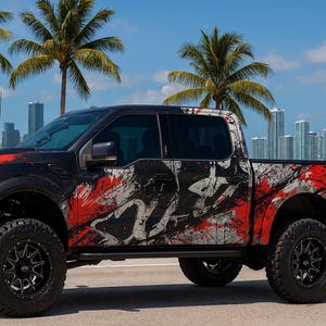 May include: Black Ford Raptor pickup truck with a custom paint job featuring red and white splatter designs. The truck has large black wheels and is parked on a road with a city skyline and palm trees in the background.