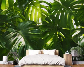 May include: A bedroom with a large, vibrant green monstera leaf wallpaper. The bed has a light wood frame and a white comforter. A small wooden side table holds a potted plant and a bottle. The room has a modern, natural aesthetic.