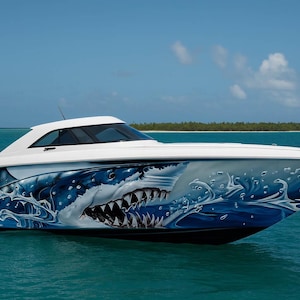 May include: A white speedboat with a shark design on its side. The graphic depicts a shark's open mouth and teeth, with water effects. The boat is on the sea under a blue sky.