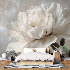 May include: A large, off-white peony flower mural is the focal point behind a bed with a light-coloured duvet. The room features a wooden bed frame, a patterned rug, and a small side table with a potted plant. The overall aesthetic is soft and neutral.