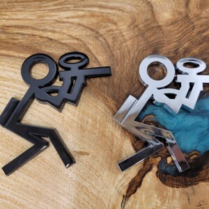 May include: Two stick figure decals, one black and one silver, are shown on a wooden surface. The figures are in a sitting position with their legs crossed.