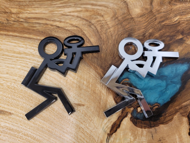 May include: Two stick figure decals, one black and one silver, are shown on a wooden surface. The figures are in a sitting position with their legs crossed.