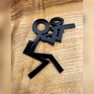 May include: Black stick figure figurine with arms raised in the air, as if celebrating. The figurine is made of a glossy material and is sitting on a wooden surface.
