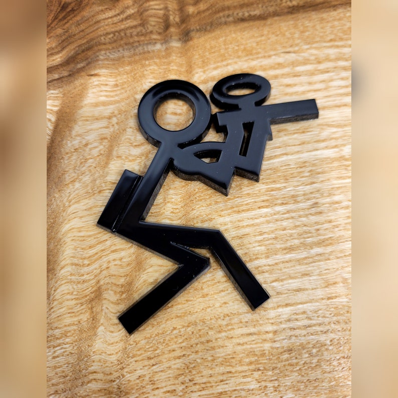 May include: Black stick figure figurine with arms raised in the air, as if celebrating. The figurine is made of a glossy material and is sitting on a wooden surface.