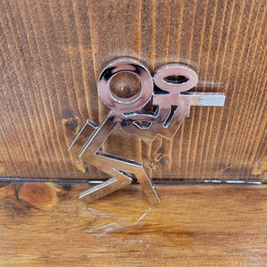 May include: A silver metal charm with the letters "K" and "O" intertwined. The charm is on a wooden surface.
