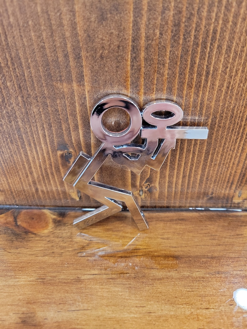 May include: A silver metal charm with the letters "K" and "O" intertwined. The charm is on a wooden surface.