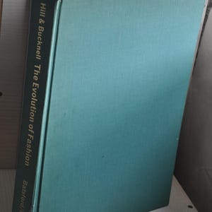 May include: A hardcover book titled "Hill & Bucknell The Evolution of Fashion" with a teal cover. The book's spine displays the title and author's name in gold lettering. The book is slightly open, revealing white pages.