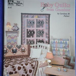 May include: A book cover titled "Baby Quilts from Grandma" by Carolann M. Palmer. The cover features a display of baby quilts with various patterns, including cat and heart designs. The quilts are in shades of pink, brown, and gray, with a white bed frame and a small table.