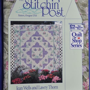 Op de afbeelding: Een boekomslag voor "The Stitchin' Post" met een quiltontwerp met een bloemen- en geometrisch patroon in tinten paars, wit en groen. De omslag bevat de tekst "Quilt Shop Series" en de namen Jean Wells en Lawry Thorn.