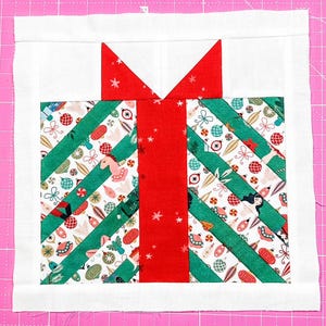 May include: A square fabric quilt block featuring a central red vertical ribbon design with a bow at the top. The background consists of diagonal strips of white fabric patterned with colorful Christmas ornaments and green solid fabric strips.