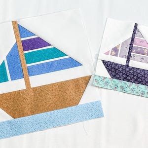 May include: Two fabric sailboat quilt blocks. One features a blue, teal, and purple striped sail and a brown hull. The other has a purple floral sail and a dark blue hull. Both are on white backgrounds.