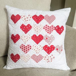 May include: White square pillow with a quilted design featuring red and white heart appliqués. The hearts have various patterns, including stripes, polka dots, and floral designs. The pillow is on a neutral-colored surface.