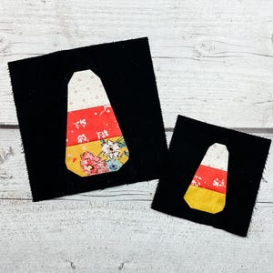 May include: Two black fabric squares each feature a candy corn design. The candy corn is made of white, red, and yellow fabric with floral patterns. The larger square is approximately 10 cm by 10 cm, and the smaller one is about 6.5 cm by 6.5 cm.